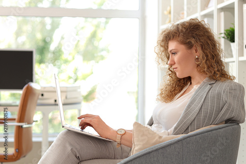 Young pretty businesswoman sitting on armchair with laptop in office