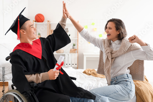 Male graduate in wheelchair with diploma and his friend giving each other hig...
