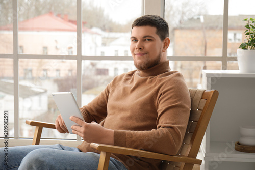 Young bearded man using tablet computer in armchair near window at home
