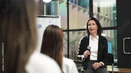 Professional businesswoman giving presentation with microphone to colleagues in modern office, sharing business ideas and strategy during corporate meeting, training and team communication.