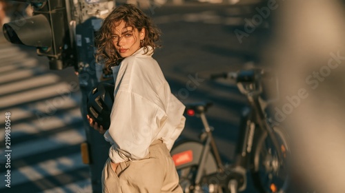 Adult woman with mixed heritage pauses at curbside, one hand holding bicycle helmet and other in pocket. Her composed expression and urban street portrait styling create polished, modern look.