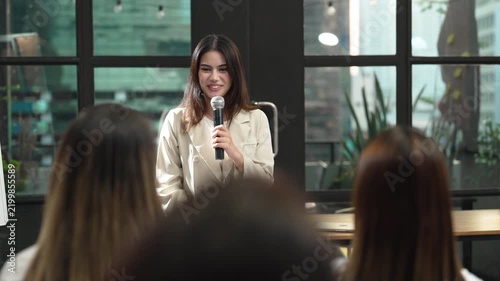 Young businesswoman giving presentation with microphone to colleagues in modern office, speaking during corporate training, team meeting and professional workplace communication event.