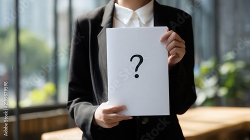 Luxury corporate photography of a professional holding a clean white document featuring one large minimalist black question mark, modern glass office