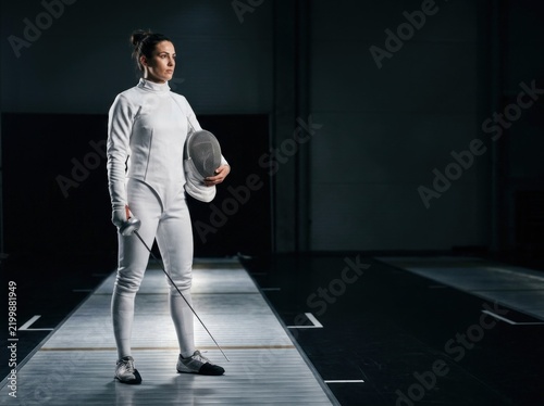 Professional fencing athlete in full white uniform holding mask standing on dark tournament strip piste