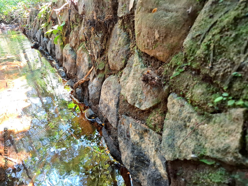 Retaining wall made of river stones with moss reflecting in clear water stream.