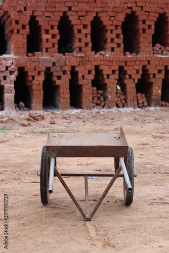Blurred Metal Wheelbarrow in Front of a Traditional Red Brick Kiln with Stacked Clay Bricks.