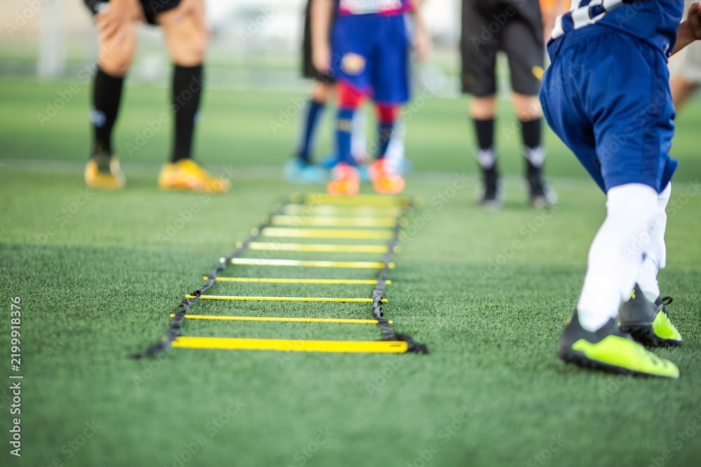 Obraz premium Selective focus of a yellow speed agility ladder placed on green artificial turf field for footwork training drills with blurred kids practicing soccer in background.