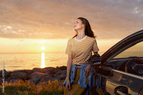 Foto Young woman stands beside a car at sunset on a rocky coast, looking up with a relaxed smile, capturing a peaceful, hopeful moment