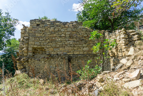 The stone wall of a ruined house. The masonry is made of light brown stones of varying sizes. The crumbled wall is visible. A sky with clouds is in the background.