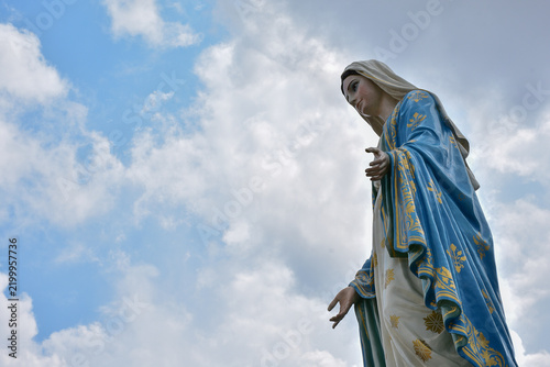 Obraz na plátně Virgin Mary statue blue white robes beneath bright cloudy sky representing Chris