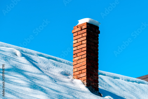 Architectural winter detail of a tall red brick chimney rising from a thick snow-covered sloped roof, topped with a perfect white snow cap and small icicle drip