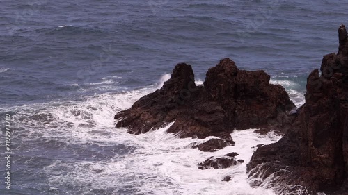 Sharp dark rocks jut out of the open sea, waves washing gently around their base under a calm sky. Jagged Rocks Standing in Open Ocean Water