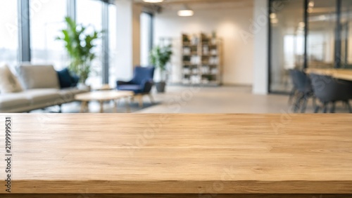 Wooden tabletop in foreground with blurred modern office lounge, sofas, chairs, plants and shelving visible in the bright interior