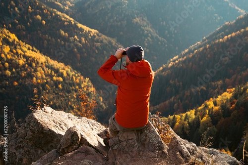 Man sitting on a rocky ledge and looking through binoculars at an autumn mountain landscape.