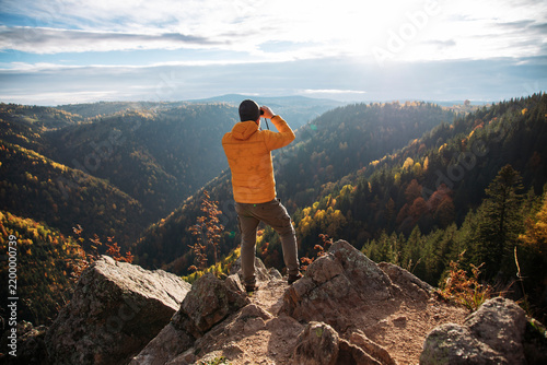 Man standing on a rocky ledge and looking through binoculars at an autumn mountain landscape.
