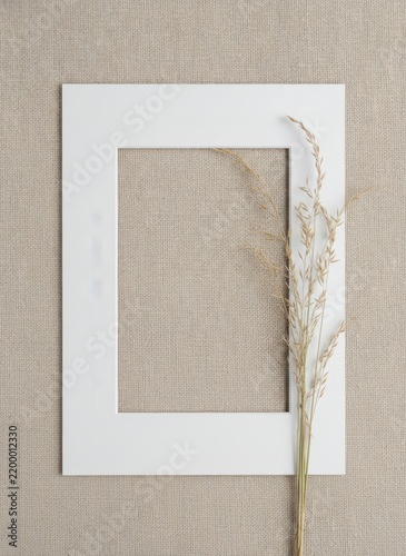 White rectangular frame mat with a single stalk of dried wild grass placed on a light beige textured linen fabric background