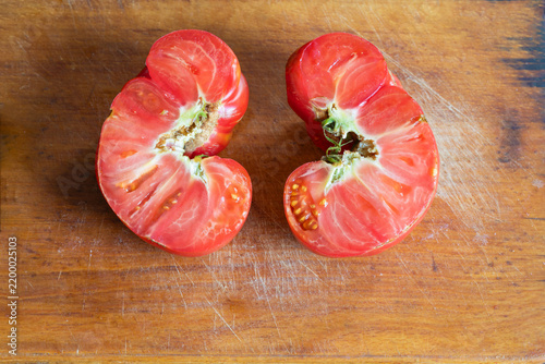Two halves of ugly, naturally misshapen red tomato lie cut open on worn wooden cutting board, revealing fleshy interior, seed chambers, and irregular structure. Natural imperfections ready for food