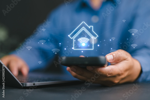 Smart Home Technology with Wireless Network Control. Man using smartphone and laptop with Wi Fi and house icons, symbolizing IoT connectivity, remote access, and modern digital lifestyle at home.