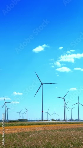 Multiple wind turbines placed in green rural farmland. White wind generators stand across green agricultural field under bright blue sky with white clouds.