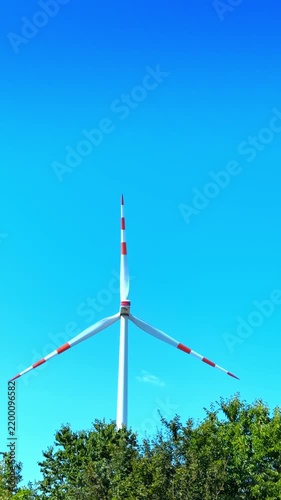 Single wind turbine rising above green tree canopy. A low angle shot of a white wind generator with red striped blades behind dense foliage under clear sky.