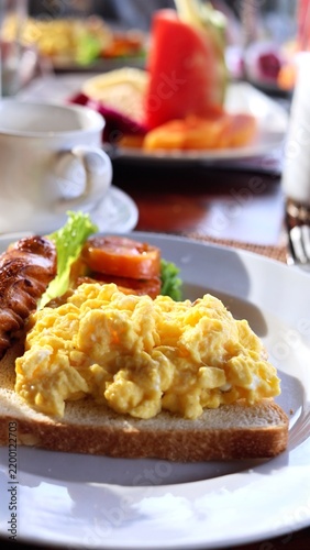 Scrambled eggs on toast dominate breakfast plate, paired with croissant and fruit. This appetizing morning meal has bright, relaxed cafe feel and simple plated presentation with lettuce and salad.