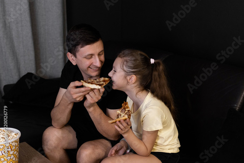 Happy father feeding his daughter a slice of delicious pizza while sitting on a leather sofa during a fun family movie night at home. Evening family entertainment concept.
