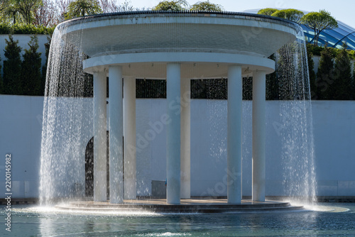 Krasnodar, Galitsky Park. Circular rotunda with cascading water flowing down its sides like curtains. Rotunda reflect in lake of love. Surrounding greenery includes bushes and trees. Cloud Park