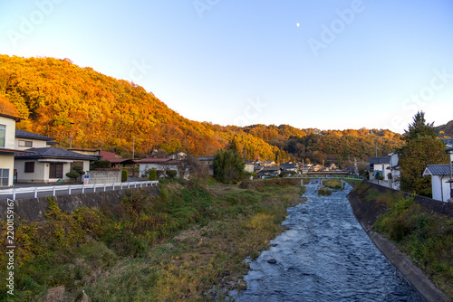 Fototapeta 夕日を受けて黄金色に輝く紅葉の山と穏やかに流れる川沿いの田舎の風景