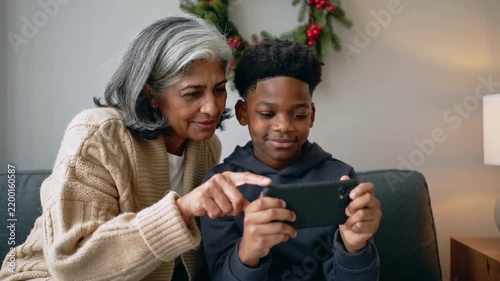 Leaning grandma wearing cardigan guiding grandson holding phone on sofa, pointing at screen