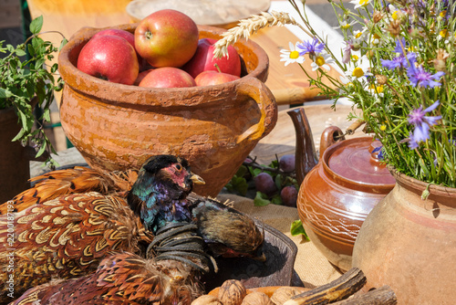 Rustic medieval market display featuring a harvested pheasant and earthenware pots filled with red apples and wild meadow flowers.