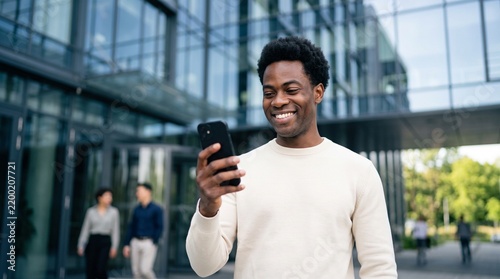 Smiling African American man using smartphone outdoor for mobile banking or digital payment app near glass office building, happy user checking fintech financial investment or online trading