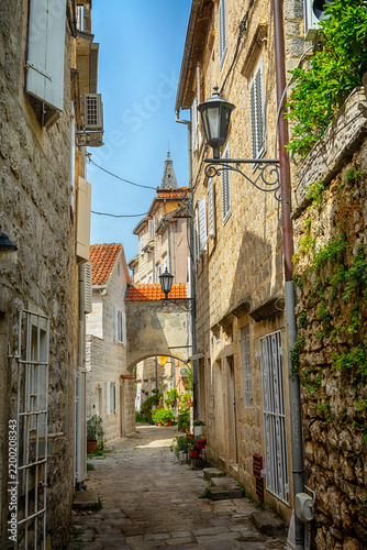 Narrow cobblestone street in Perast, Montenegro. Stone buildings with red roofs line the path. A vintage street lamp hangs above, creating a charming atmosphere.
