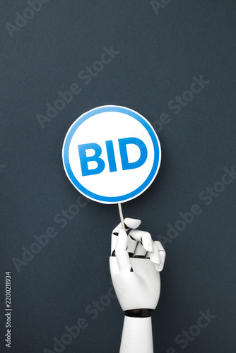 Foto A white robotic hand holding a blue bidding paddle with BID signs on a dark gray background