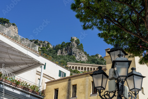 Ancient Torre dello Ziro watchtower on a cliff in Amalfi Italy. Ornate street lamps, colorful balconies, Mediterranean buildings, rocky cliffs, and deep blue sky in scenic European hillside town
