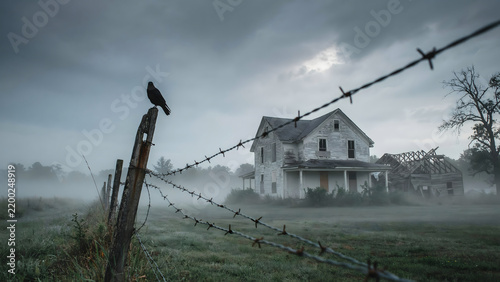 A black crow perched on a wooden fence post with barbed wire in front of a derelict, abandoned house under a dark, gloomy, and foggy sky at dusk