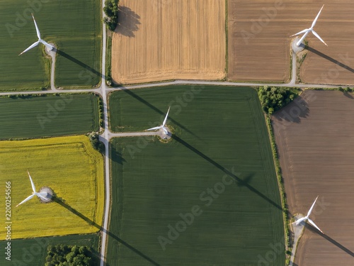 Top-down view of wind turbines casting long geometric shadows across cultivated farmland and narrow service roads.