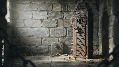 Medieval iron maiden torture device with sharp spikes and a padlock, standing inside a dark stone dungeon cell with chains and scattered bones