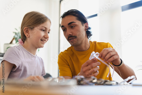 A young man and a smiling young girl sit together at a table, carefully choosing new eyeglasses. He holds up a pair for her to examine during their optician appointment.