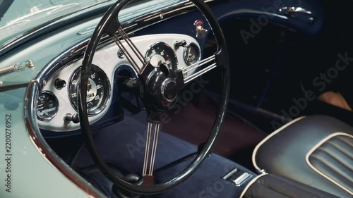 Close-up view of a vintage car's black steering wheel and white instrument panel with chrome gauges inside the classic vehicle cabin during daytime.