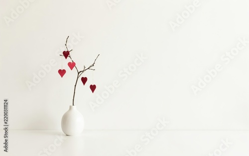 White vase with bare branches adorned with red hearts against a plain background, decorations