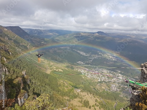 Un homme en train de « slacker » sur une Highline entre deux falaises, avec un arc-en-ciel en toile de fond (Rocher de Rochefort, causse Méjean, 