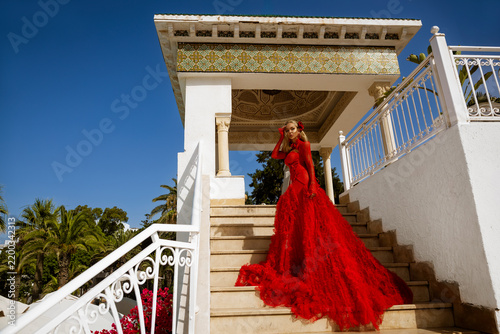 Glamorous Blonde Woman in a Voluminous Red Tiered Gown Posing Dynamically on Marble Stairs by a Luxurious Tropical Resort Pool with Palm Trees and a Bright Blue Sky