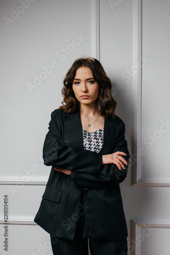 A confident young woman wearing a black blazer and tiara stands with crossed arms against an elegant white wall, blending modern sophistication with regal charm