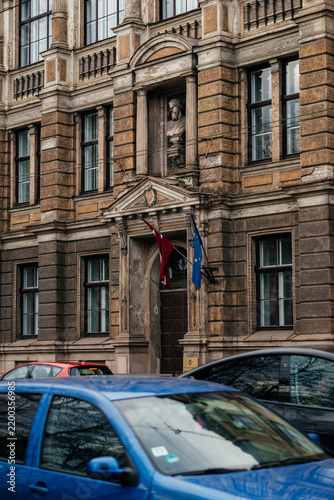 An elegant historic building with classical architecture, featuring decorative columns, arched windows, and a stone bust above the entrance, adorned with Latvian and European Union flags