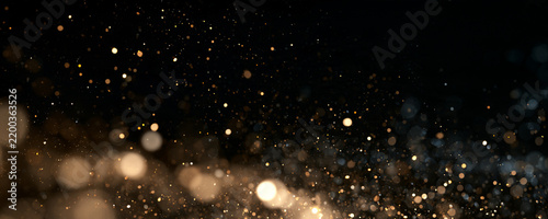 Gold dust particles scatter in a dark space. The tiny specks glow with soft warm light and varying intensity. This image captures a sparkling bokeh effect perfect for festive or background.