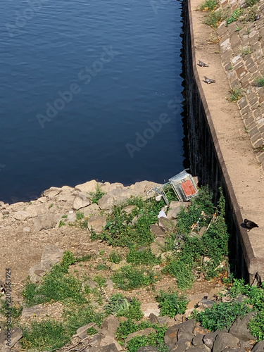 Low river water exposes a dry rocky bank with discarded shopping cart and litter beside an urban embankment, illustrating drought, pollution and environmental impact.