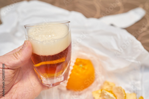 A hand holds a glass of dark amber beer with thick foam against a blurred background of a peeled orange. Ideal for travel lifestyle blogs, hospitality editorials, and beverage marketing campaigns.