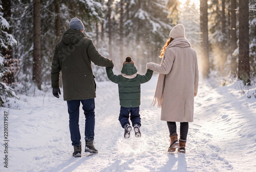 Jeune famille vue de dos en tenues d’hiver chaudes, parkas, manteau, bonnets et bottes, parents tenant les mains de leur petit garçon qui saute joyeusement dans la neige