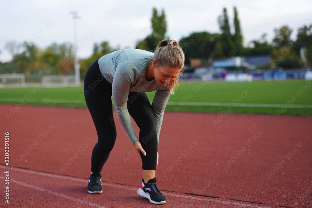 Obraz premium Female runner experiencing intense leg pain and injury on a red track, stopping mid-workout due to a muscle cramp or strain while exercising outdoors