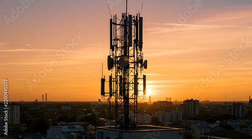 Communication mast atop the city's skyline illuminates as the sun sets. Antennas broadcast signals across the urban terrain. Skies and surrounding buildings glow with the golden hues of twilight. Mode